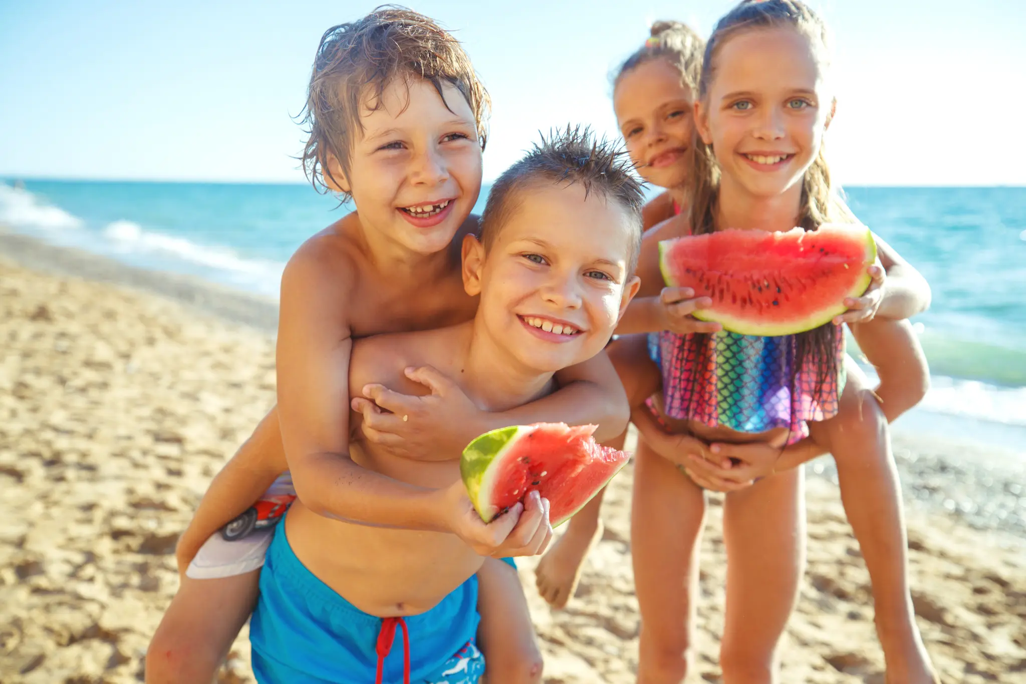 Four children on a beach smiling and eating watermelon.