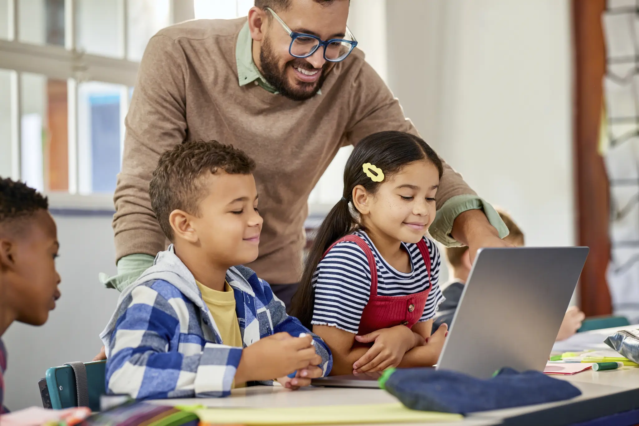 A male teacher standing behind two students while they look at a laptop in a classroom.