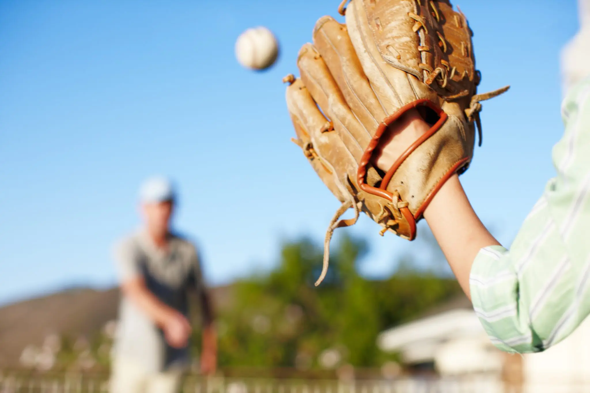 An arm with a baseball glove on the hand catching a baseball in the foreground with a person in the background.