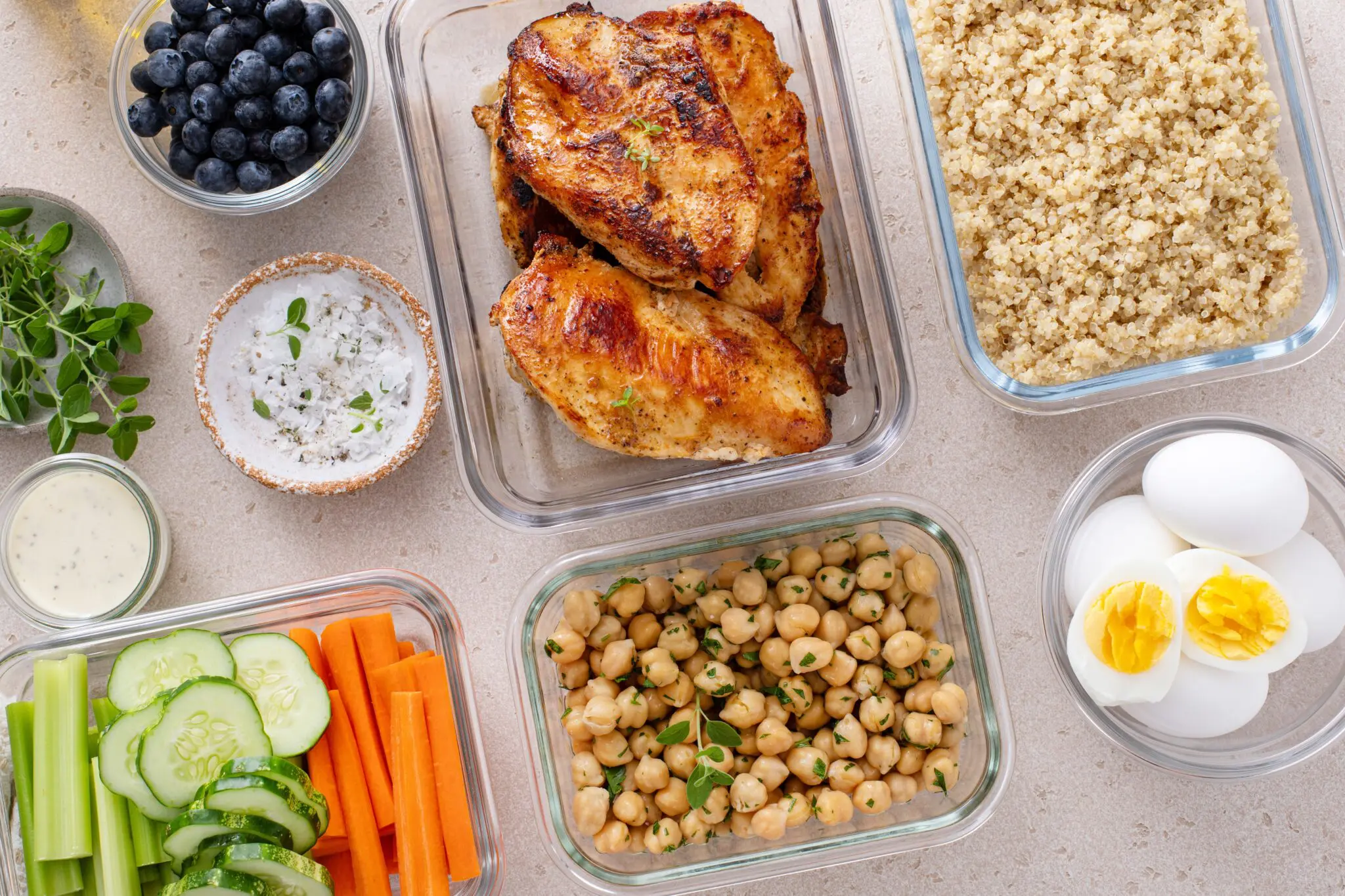 Multiple glass containers of food including chicken, grains, raw vegetables, blueberries, chickpeas and boiled eggs.