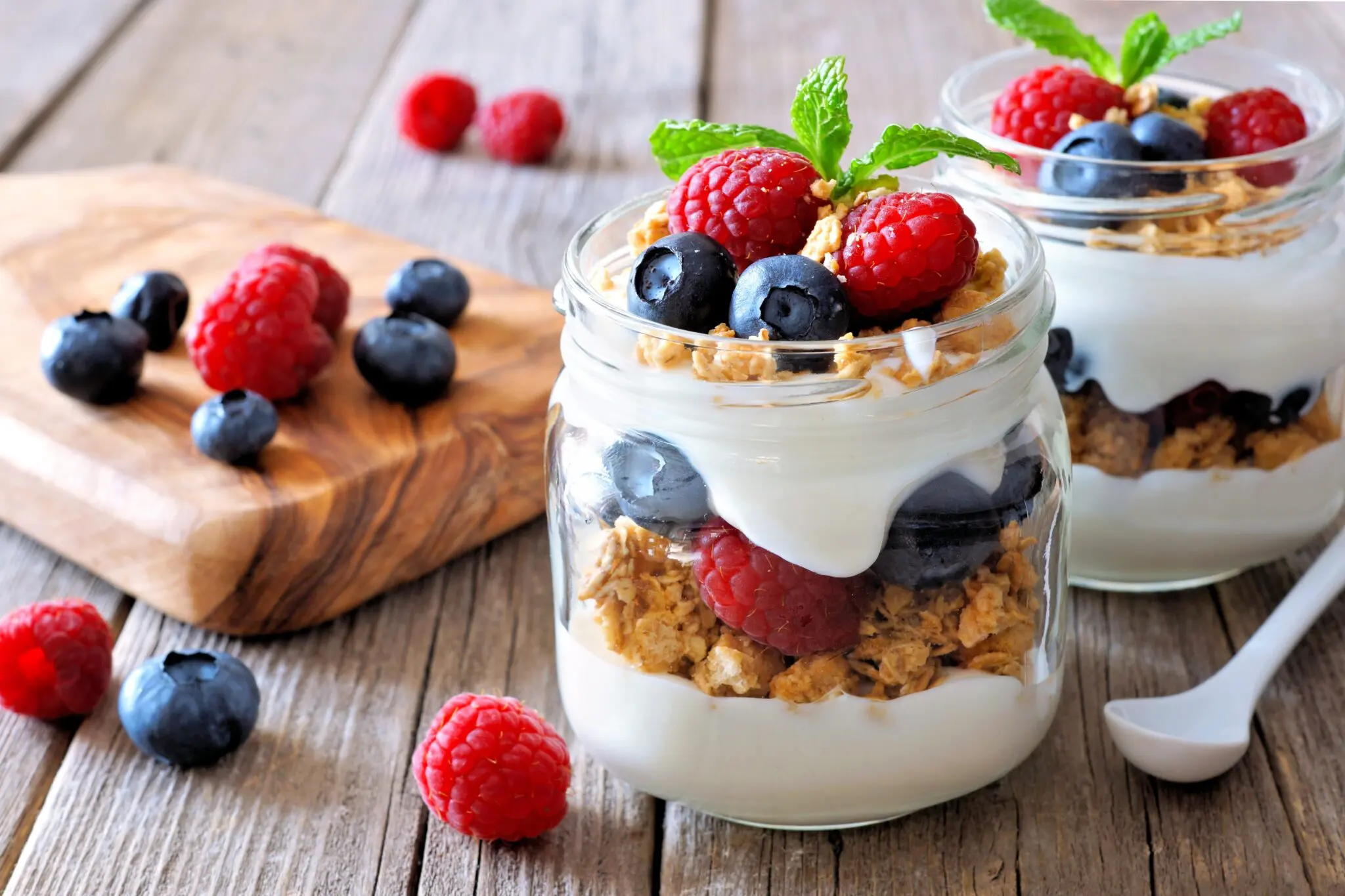 Glass jars with layers of white yogurt, granola, blueberries and raspberries in the foreground with fruit scattered around the background.