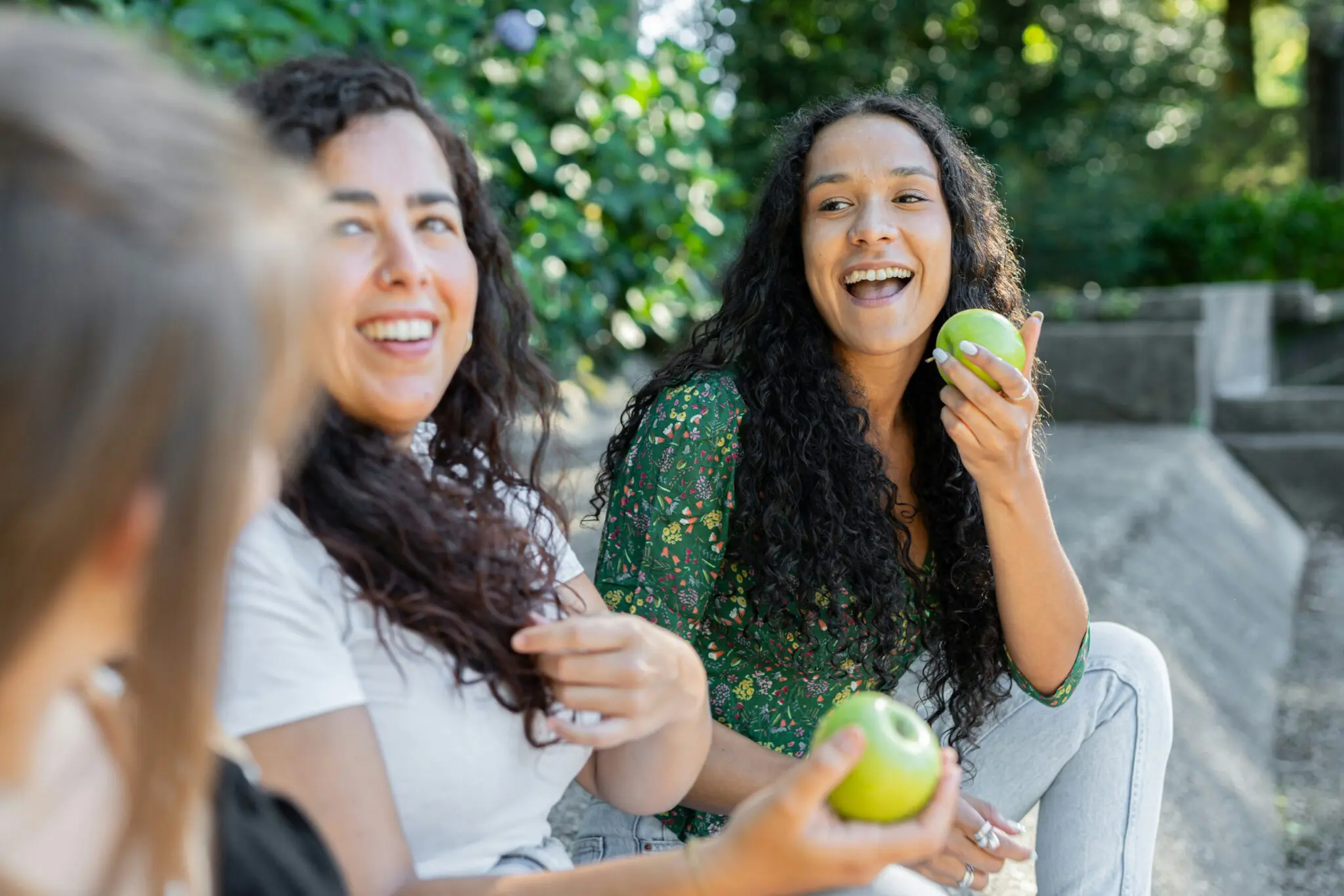 Three adult women sitting outdoors with two women facing the camera smiling and holding green apples.