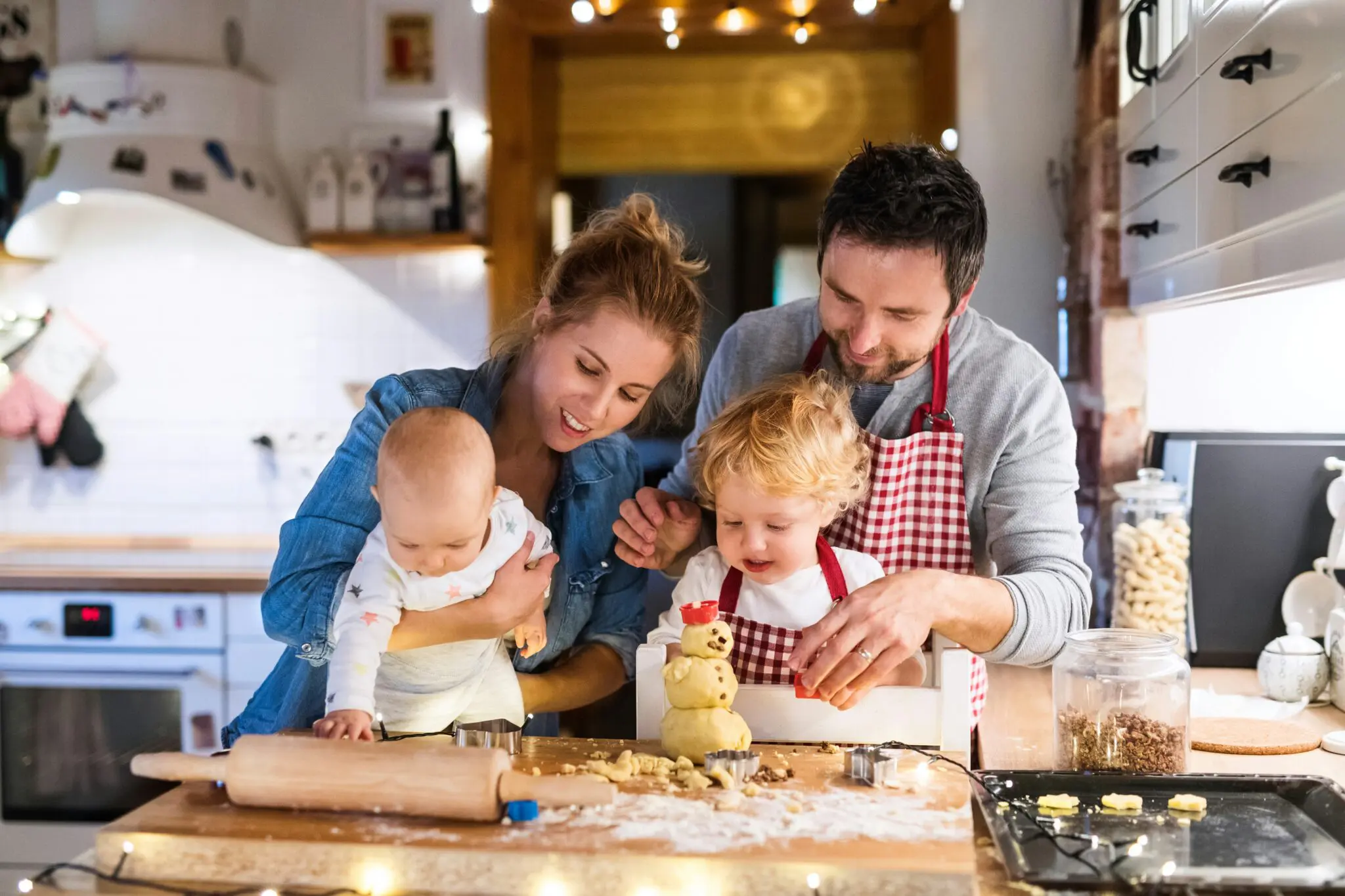 Two adults standing behind their two small children in the kitchen making cookies for the holiday.