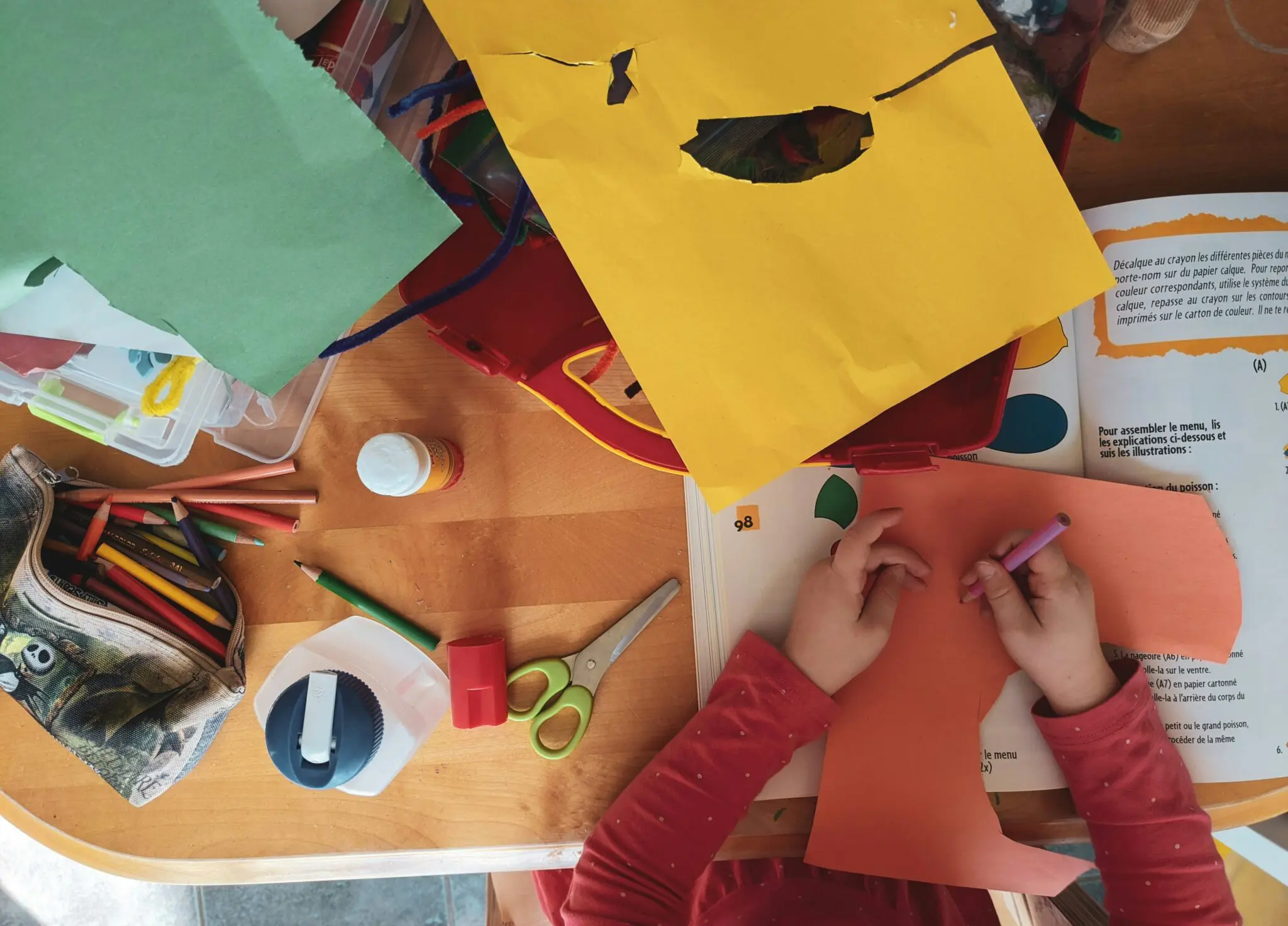 An overhead angle image of a child doing creative crafts using colored construction paper, colored pencils and other art supplies.