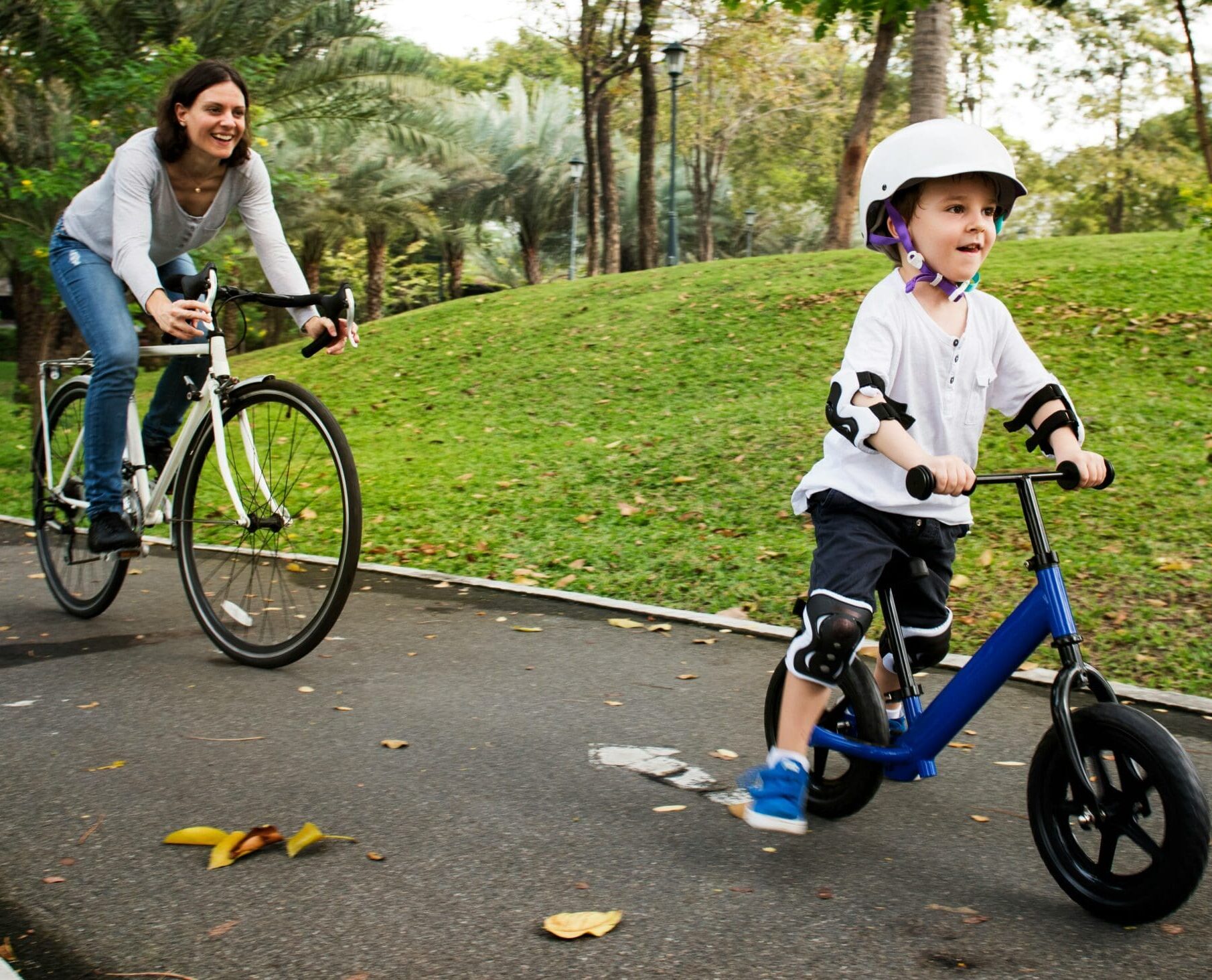 A small child wearing a helmet while riding a bicycle in front of an adult woman riding a bicycle in a park setting.