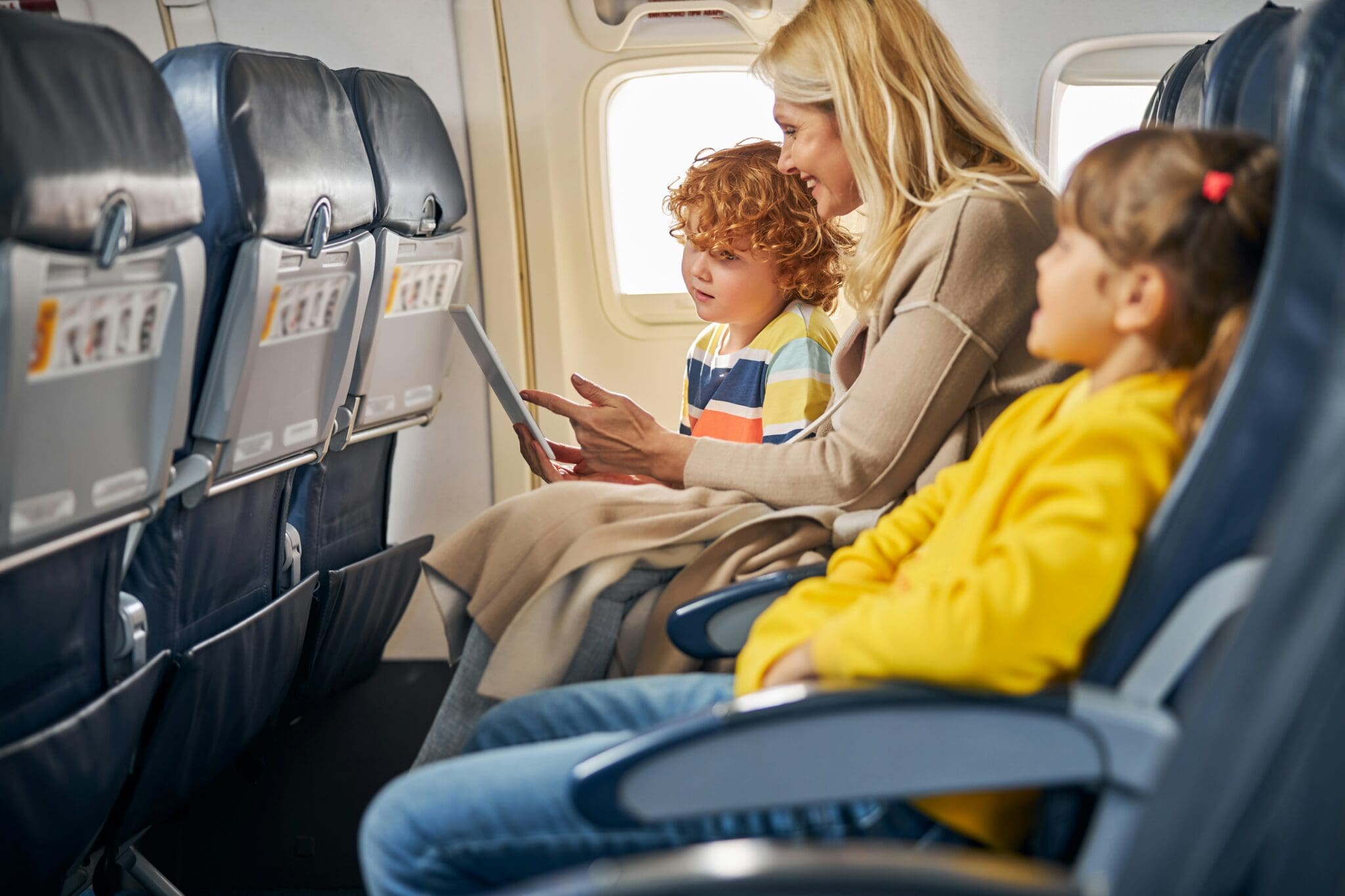 Family Vacation Two children and an adult woman seated on an airplane holding a tablet and smiling.