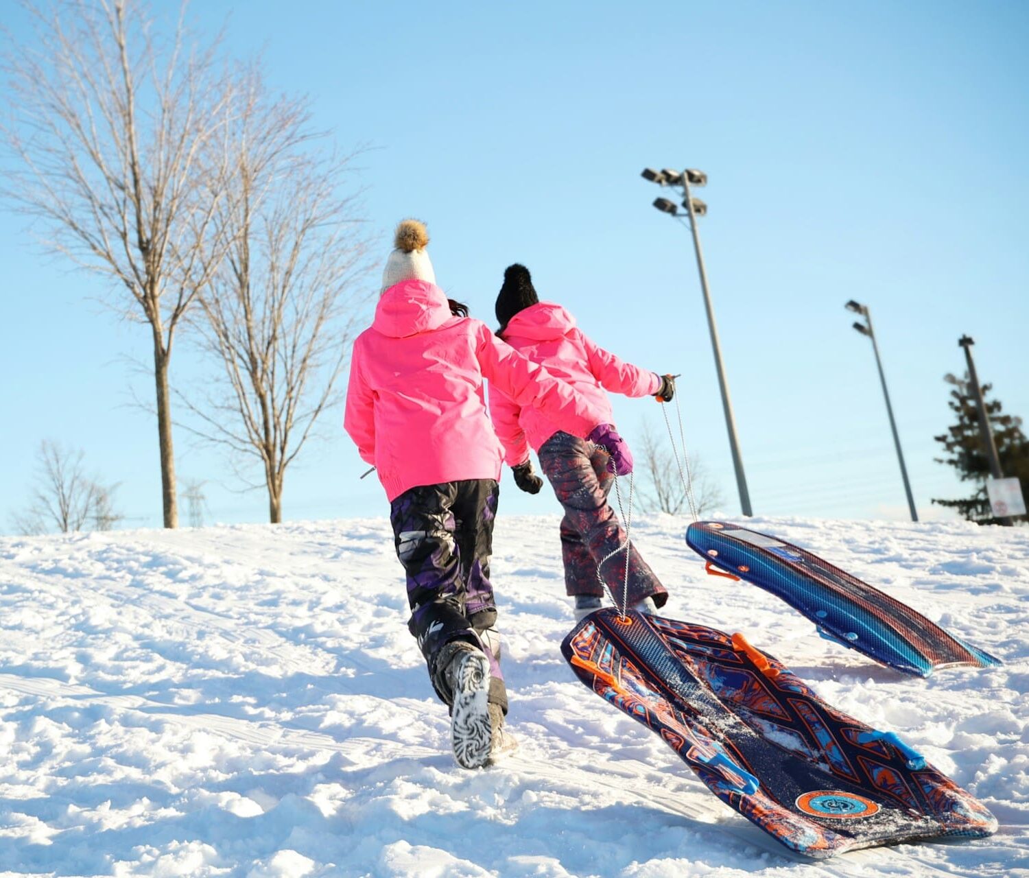 Two people in pink winter coats and hats walking up a snow covered hill pulling snow sleds behind them.